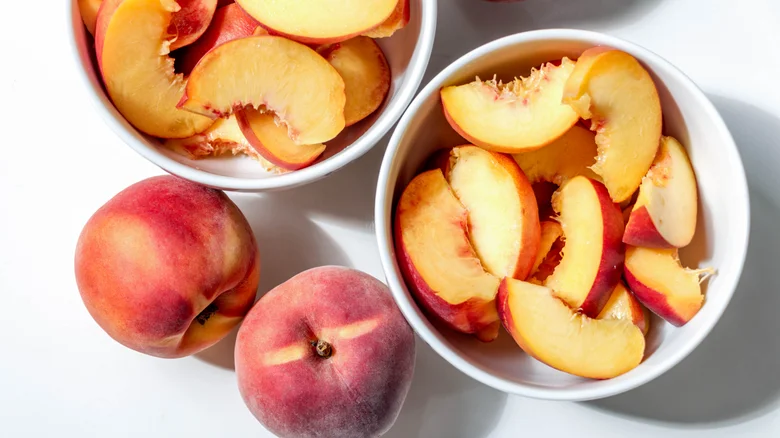 Whole ripe peaches next to bowls of sliced peaches