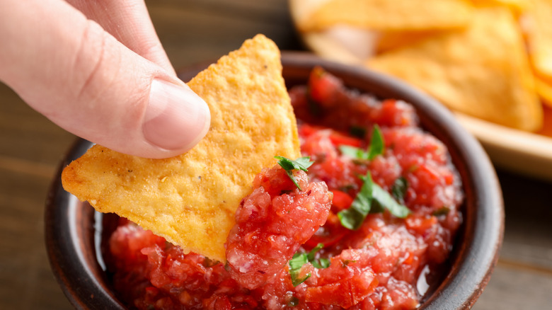 Person dipping nacho chip into spicy salsa sauce at table