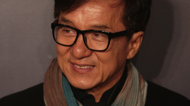 Slight high angle, looking down at Jackie Chan in shadowy lighting in front of a gray background.