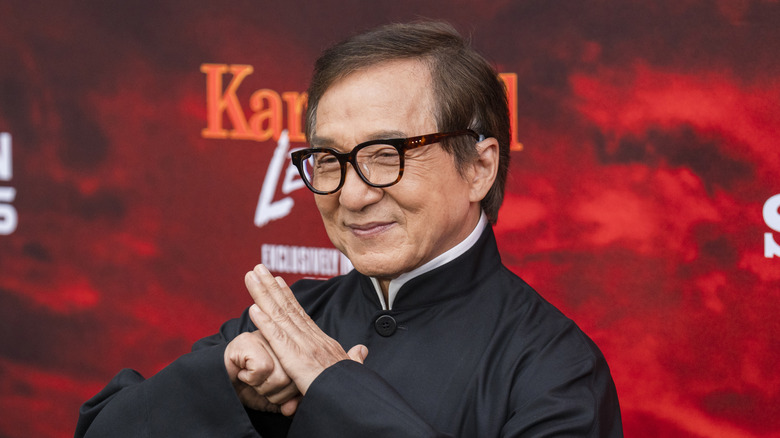 Jackie Chan in front of a red background with his hands together.