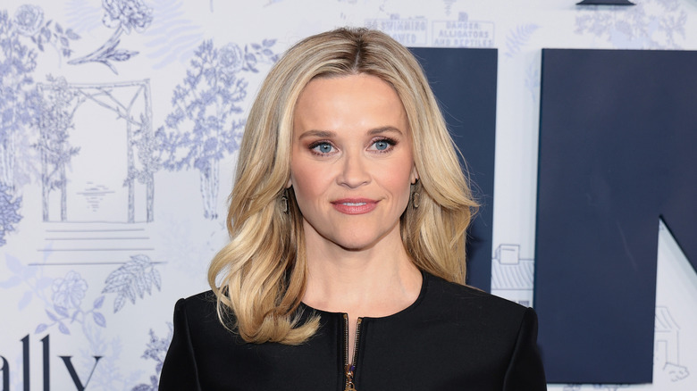 Reese Witherspoon in a black dress looking off center in front of a white background with blue details.
