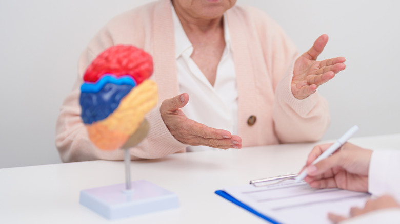 A colorful model of a brain on a stick with a woman in a pink sweater gesturing before a man writing something down.