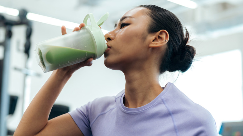 A woman drinking a green beverage in a cup inside a gym.