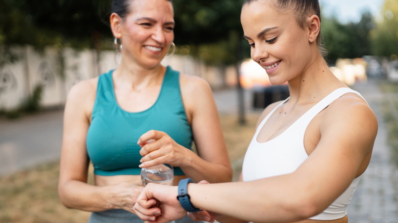 Two women standing together outside in workout clothes, one woman holds a water bottle while the other checks her watch.