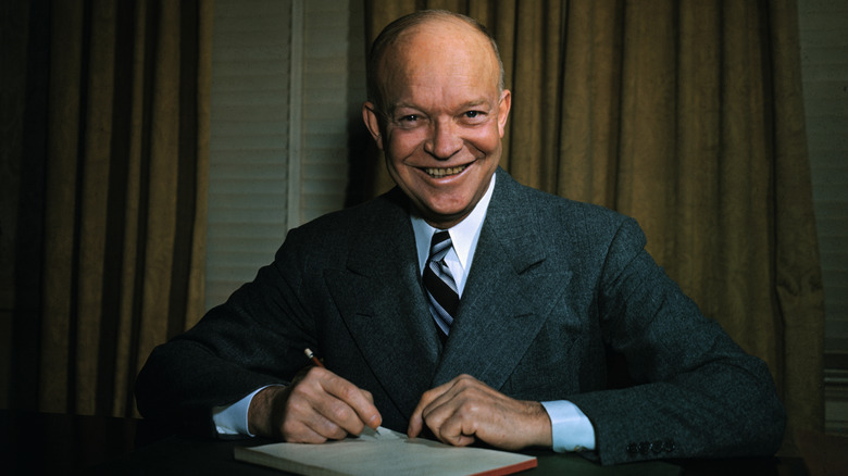 Dwight Eisenhower sitting in a suit behind a des with a pen in hand and a large smile on his face.