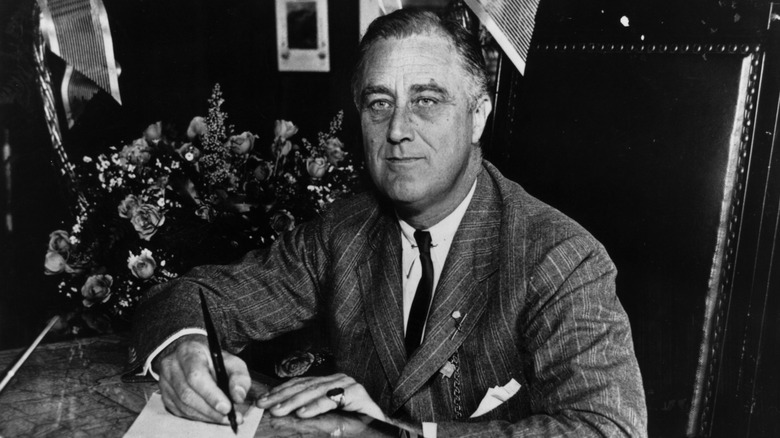 Franklin Roosevelt sitting in a chair with a pen in hand, looking off to the left side.
