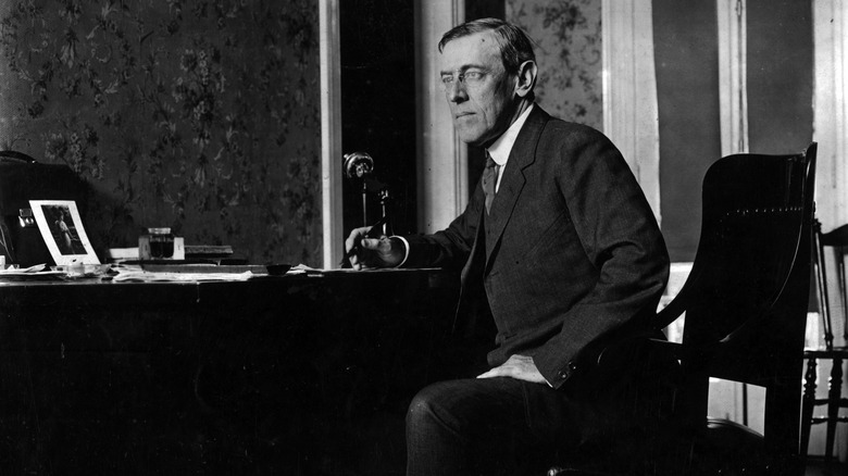 Woodrow Wilson in black and white, leaning forward over a desk with a pen in hand.