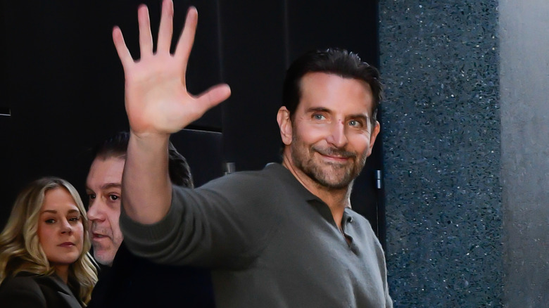 Bradley Cooper waving and smiling as he walks down a crowded street