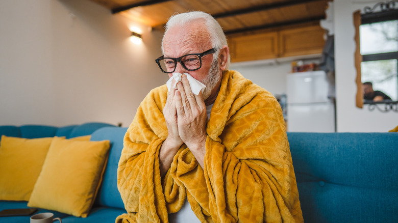 older man with cold on couch at home blowing nose