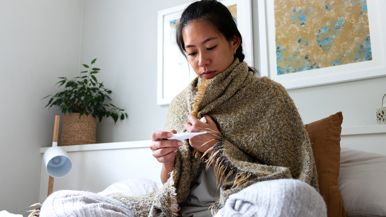 A woman wrapped in a blanket sits up in bed looking at a thermometer