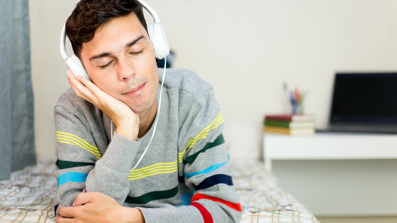 Young man lying on bed on his stomach with his head in his hand, with headphones on and eyes closed