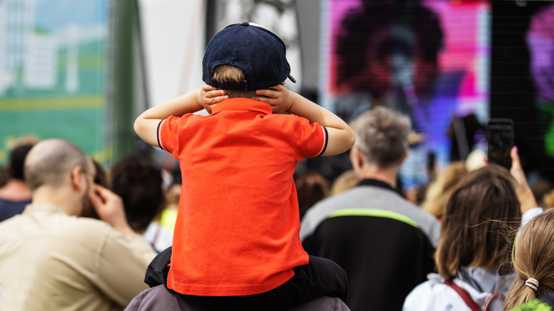 Child in an orange shirt sitting on a parent's shoulders during a concert, covering their ears