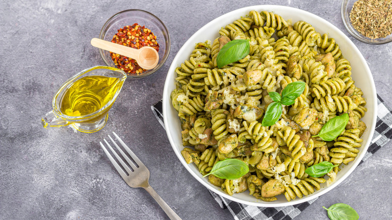 A bowl of pesto pasta with chicken topped with fresh basil and a side of olive oil and pepper flakes