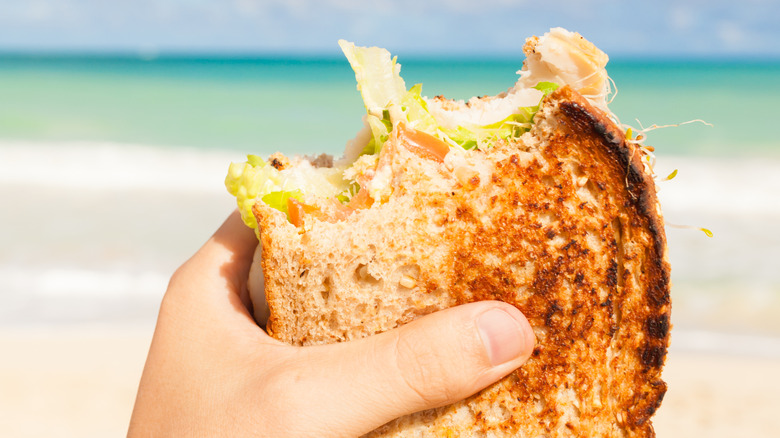 A person's hand holding a partially eaten sandwich on the beach