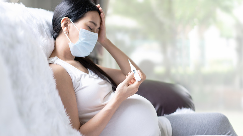 Sick pregnant woman wearing mask and holding thermometer