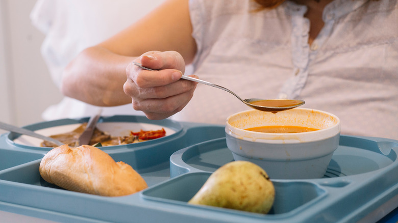 Woman in hospital bed lifting a spoon from a bowl of soup on a lunch tray