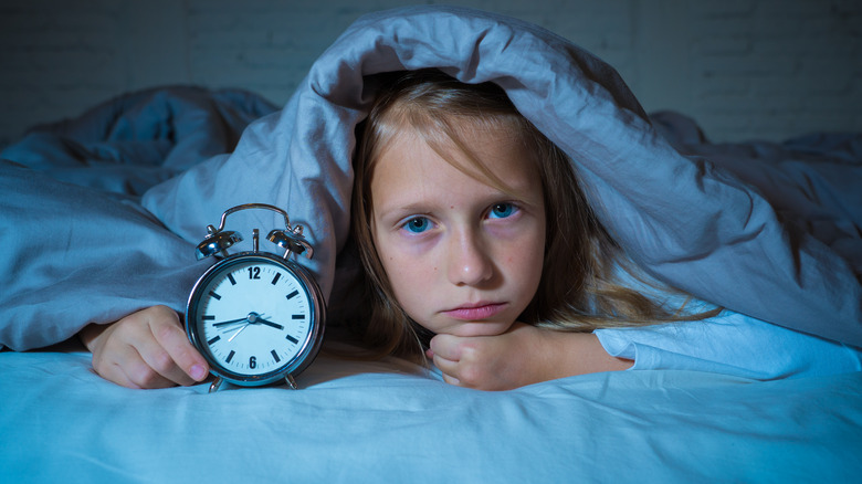 Young girl awake at night holding alarm clock under covers