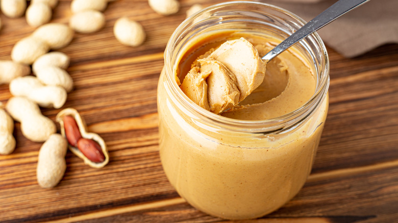 Spoon dipped in jar of peanut butter surrounded by peanuts