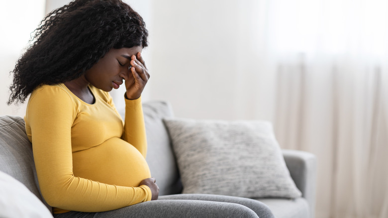 Pregnant woman sits on couch with one hand on belly and one hand on head in pain