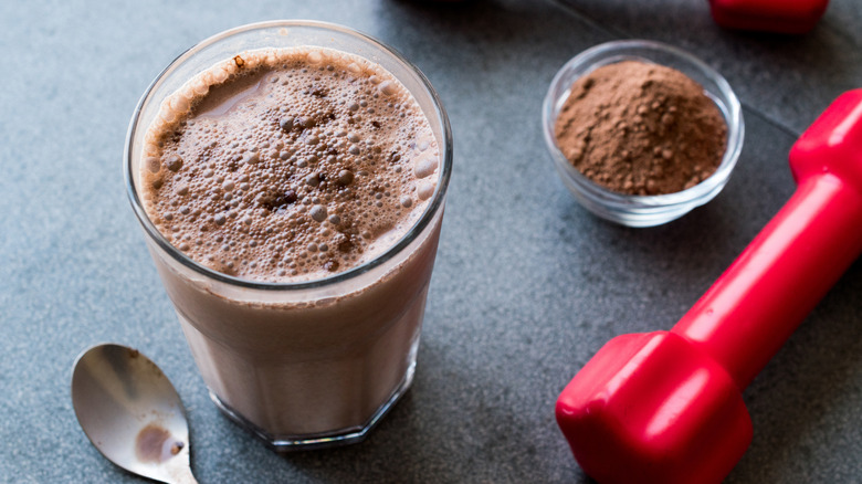 A protein shake and protein powder next to a set of dumbbells 