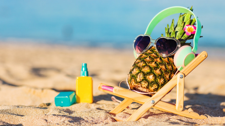 A pineapple with sunglasses and headphones reclining in a miniature lounge chair on a beach