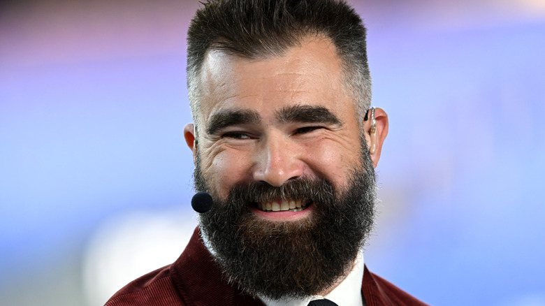 Jason Kelce smiling, a microphone by his mouth, in a red suit.