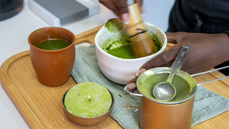 A cutting board with tools for prepping matcha laid out and used while hands whisk together the substance.
