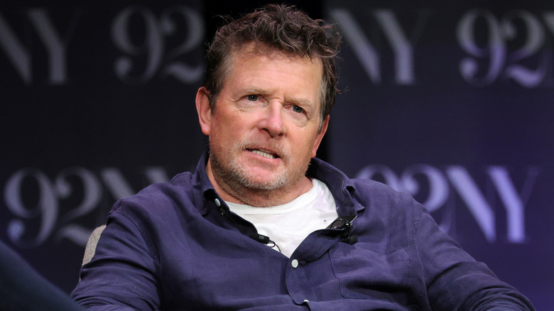 Michael J. Fox sitting while wearing a blue shirt with a white undershirt