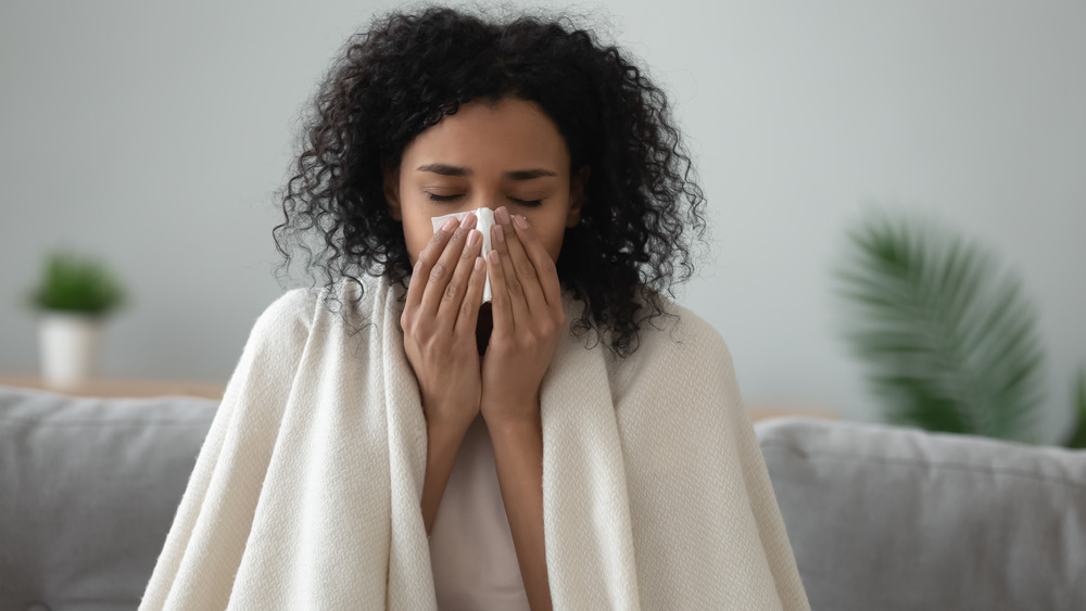 Young woman sitting on couch in blanket blowing nose