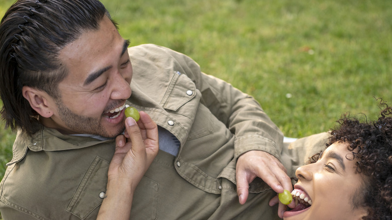 young couple smiling and feeding each other grapes while lying on the ground outside