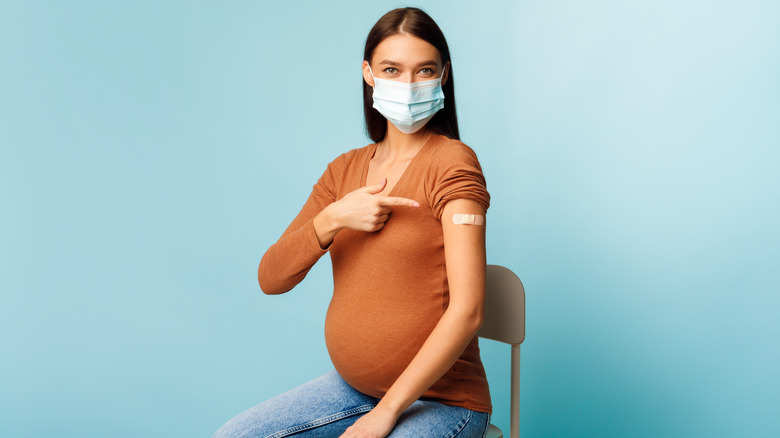 Pregnant woman with face mask on sitting in chair pointing to bandage on upper arm 