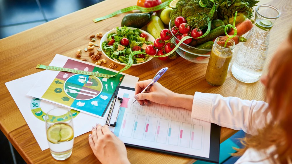 Woman dietitian in medical uniform with tape measure working on a diet plan sitting with different healthy food ingredients in the green office on background. Weight loss and right nutrition concept. Reverse dieting. 