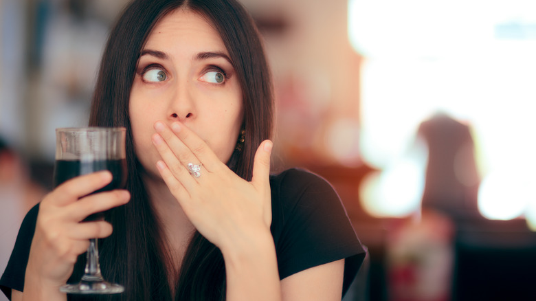 Woman covering her mouth with her left hand while holding a dark beverage in a wine glass in her right hand