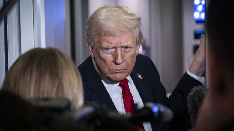 President Donald Trump on Air Force One, surrounded by reporters and leaning into a microphone.