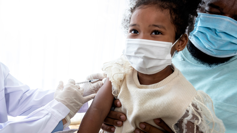 Young 5-year-old girl sitting on her dad's lap wearing masks and receiving vaccine