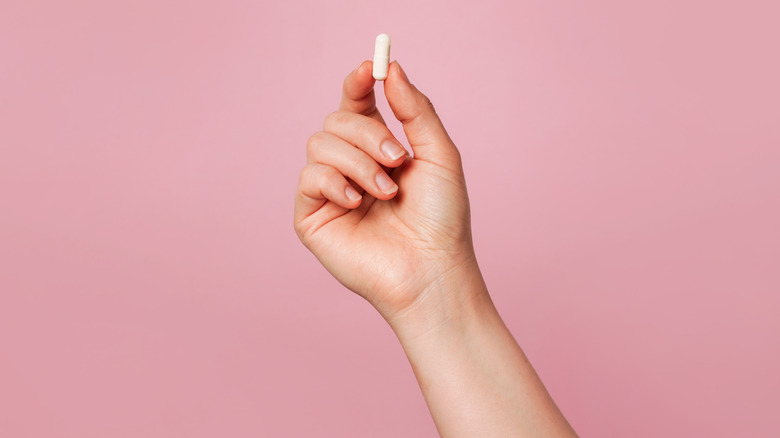 A pink background is broken by a single white hand that holds up a small white pill.