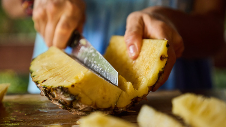 Hands cutting up a pineapple on a cutting board.