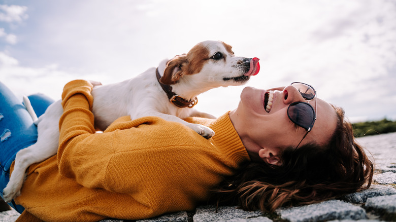 a woman lies on the ground outside laughing as her dog licks her face