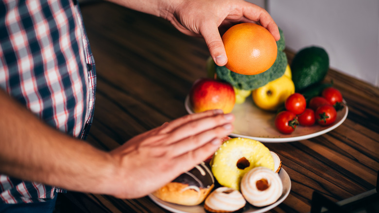 Tight shot of a person choosing an orange from a bowl while holding out a hand saying no to donuts