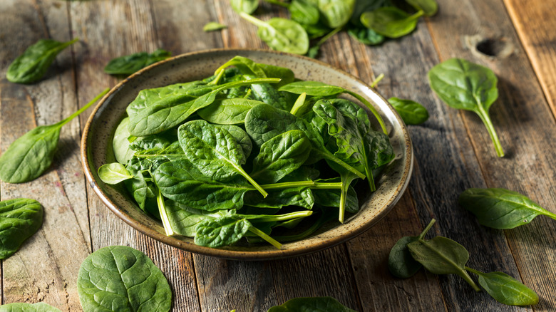 A bowl filled with spinach surrounded by spinach leaves on a wooden surface