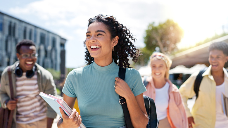 A smiling Gen Z woman holding books, followed by her friends