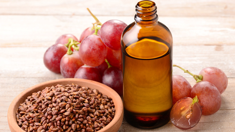 Brown bottle of grapeseed oil surrounded by red grapes and  and a bowl of grape seeds on a wooden board