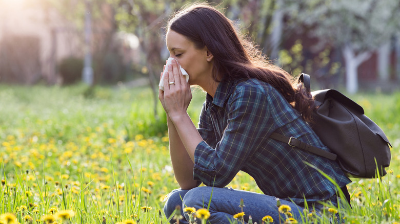 Woman wearing a backpack and blowing her nose into a tissue while crouching in a field of grass