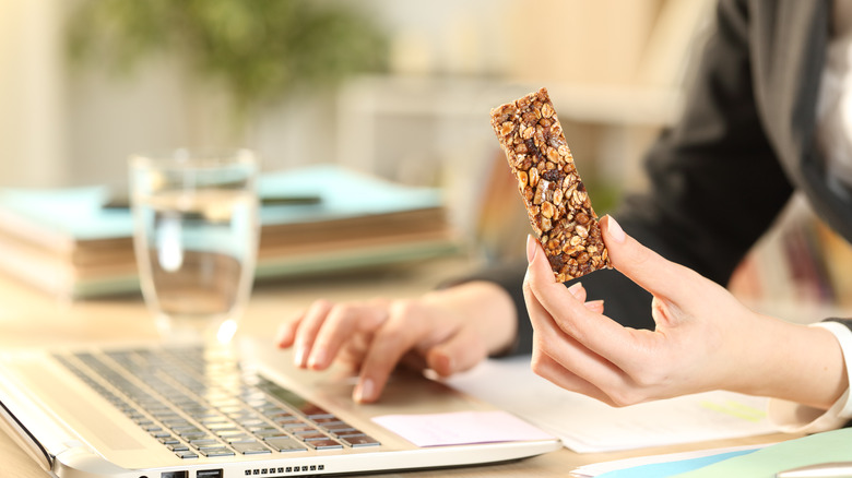 Woman holds a granola bar by her computer