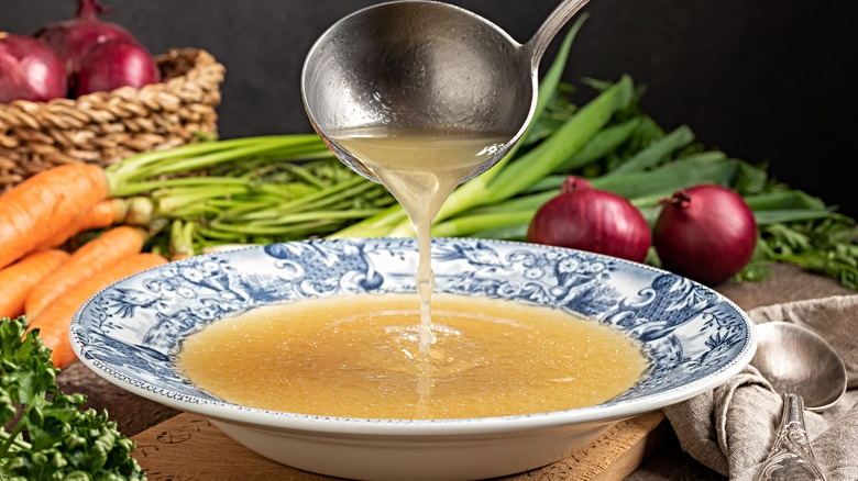 Ladle pouring chicken soup into a bowl with vegetables in the background