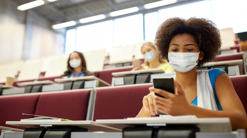 Young woman in college classroom wears face mask and checks phone