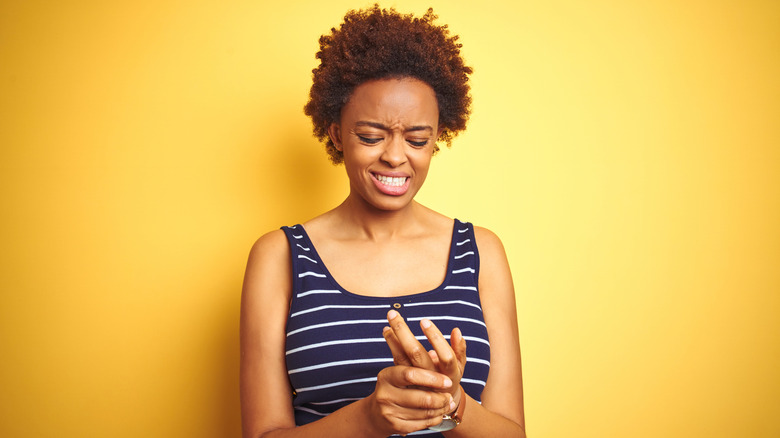 Woman in blue and white striped shirt standing in front of a yellow background holding her hand with her other hand with a pained look on her face