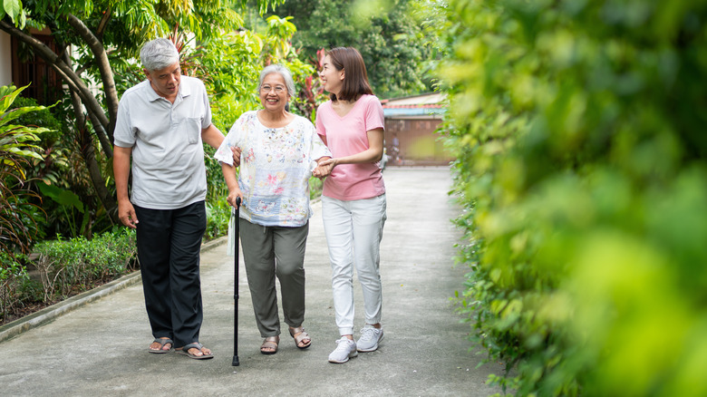 Three family members go for a walk in a garden, one walking with a cane and the other two on either side of her holding her arms