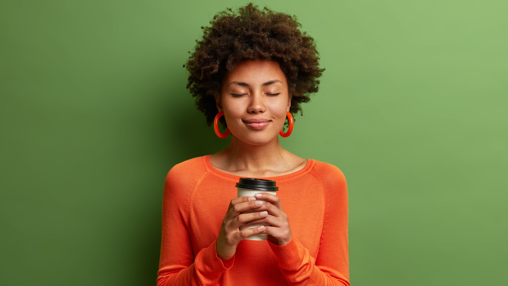 Person with short brown hair and orange top holding a cup of coffee in front of a green background