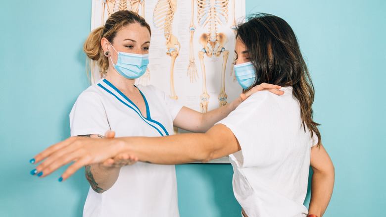 Masked doctor doing physical therapy on masked patient's arm, helping them stretch